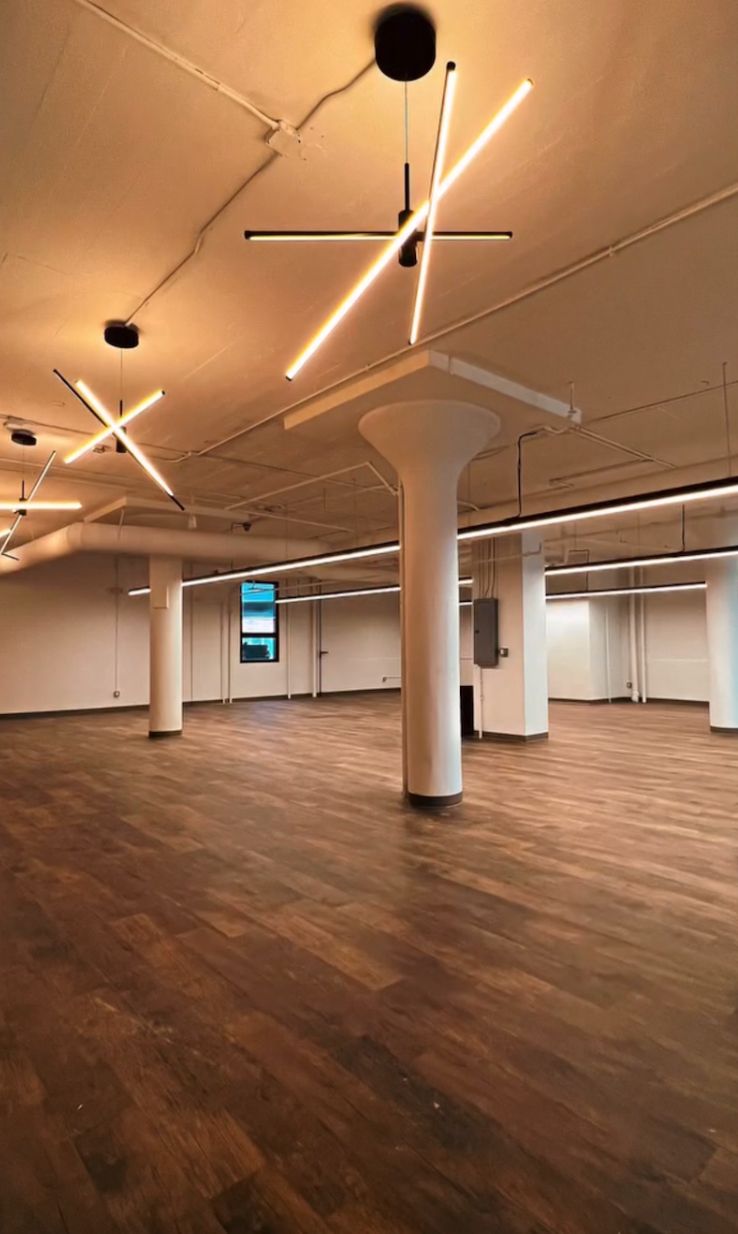 An open-plan office space featuring light wood-look flooring, white pillars, and geometric LED pendant lighting.