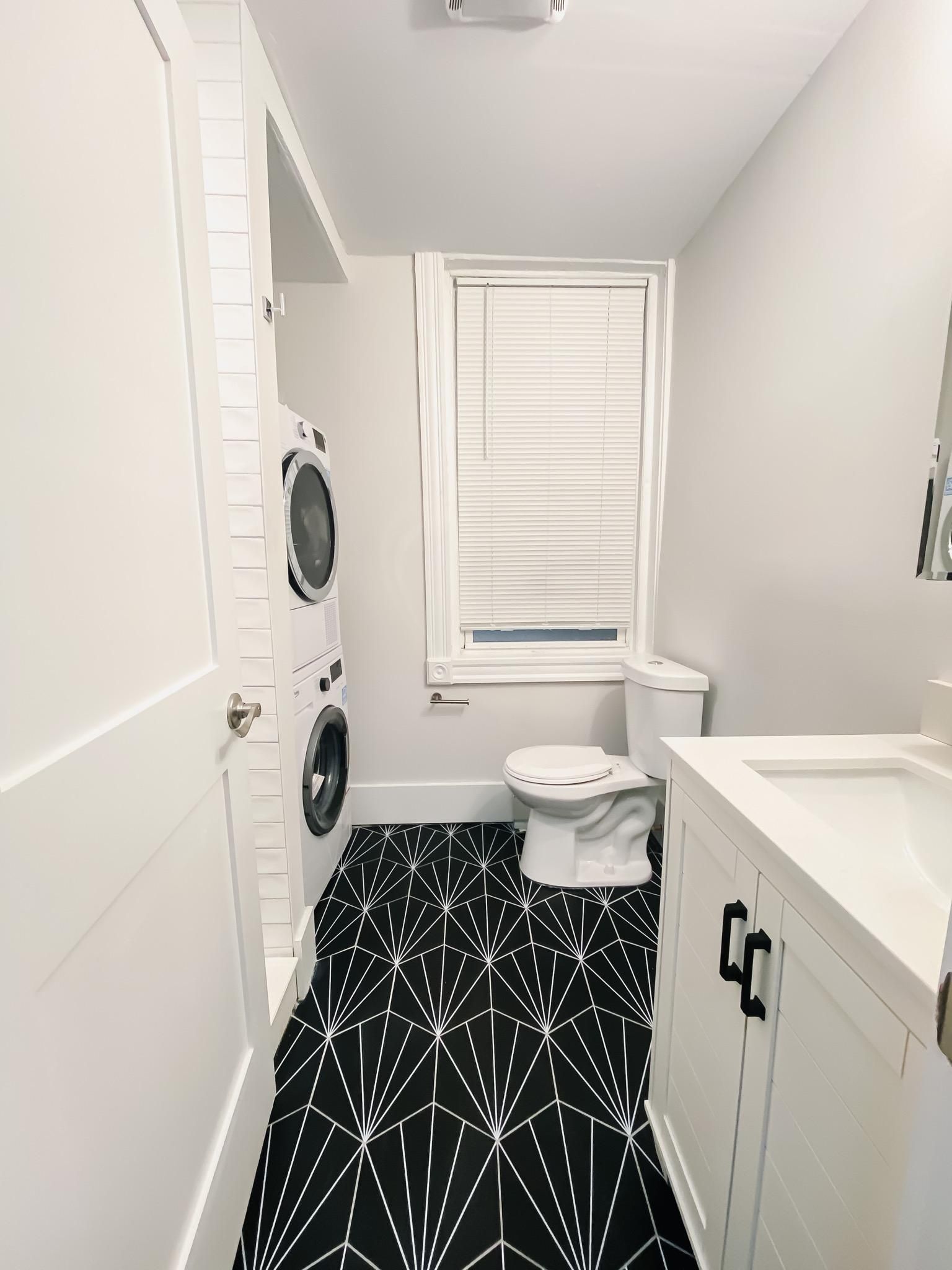 Small bathroom with a white vanity, a toilet, and a stacked washer and dryer on a black and white geometric patterned floor.