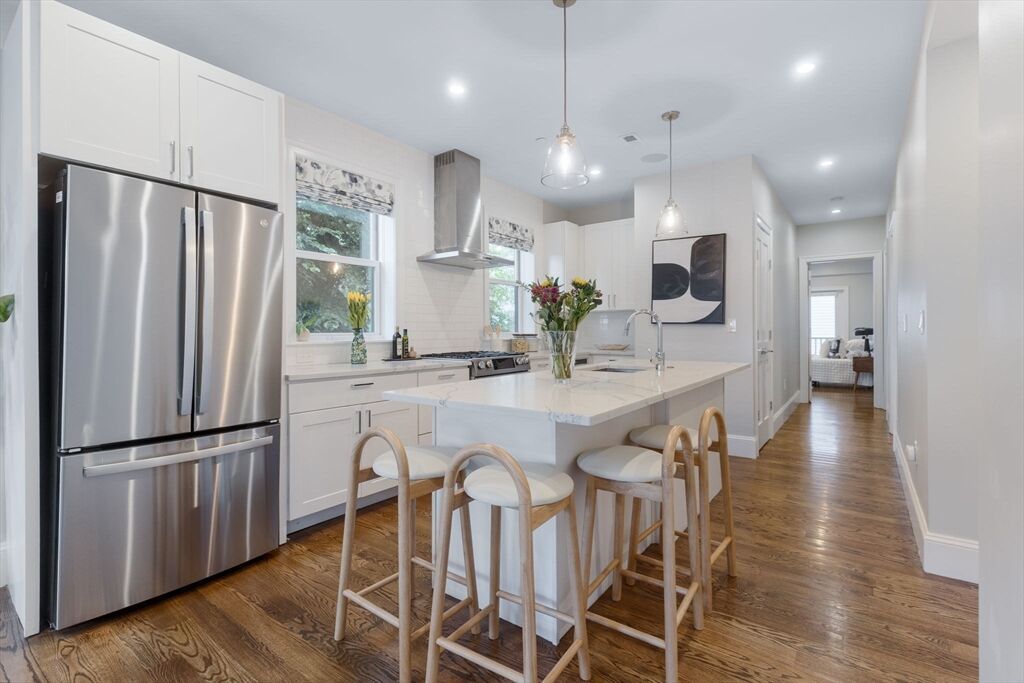 A bright, modern kitchen featuring white cabinets, a large island with three wooden stools, and stainless steel appliances.
