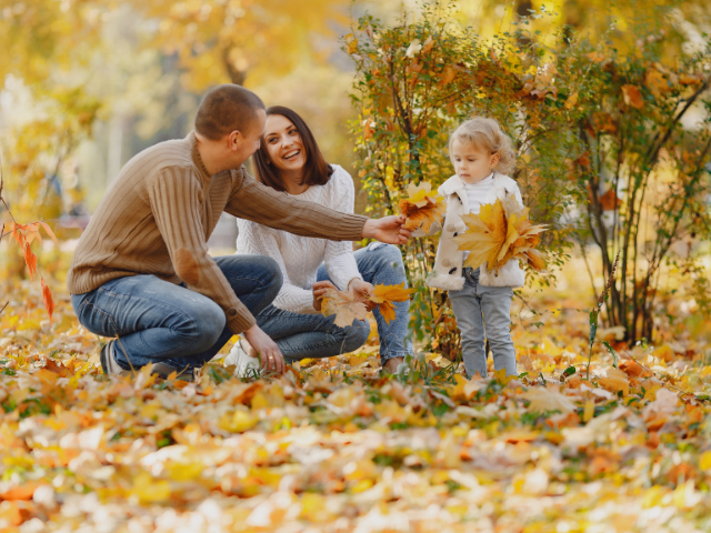 A family of three enjoys a fall day outdoors. The father crouches down handing autumn leaves to his 