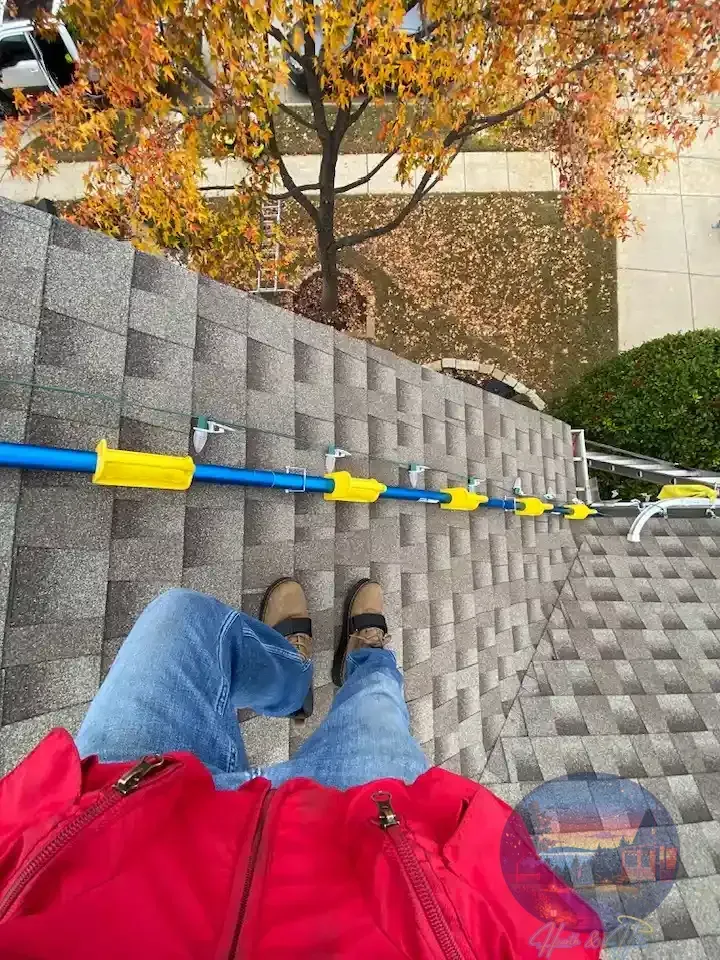 Person in red jacket and jeans on a rooftop; blue safety rail attached to shingles; fall foliage in background.