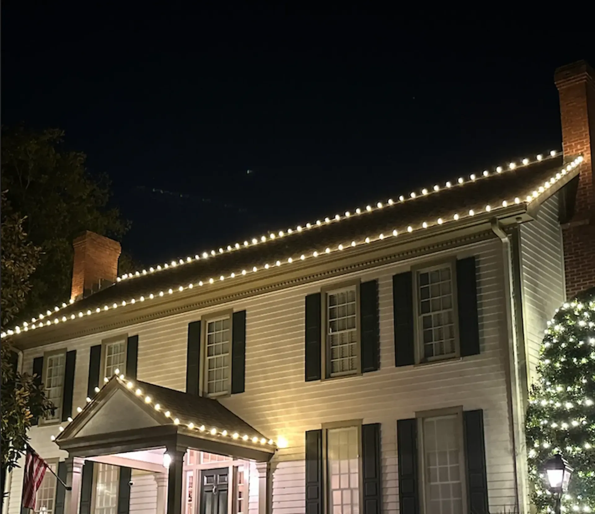 Two-story white house with lit Christmas lights along roof lines, dark shutters, and a dark night sky.