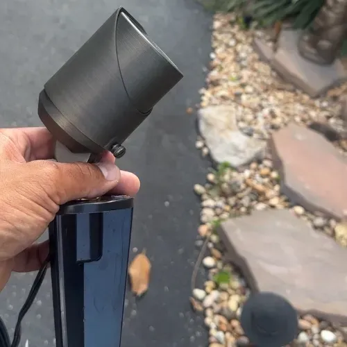 A hand holding a brass outdoor spotlight in front of a stone and gravel yard.