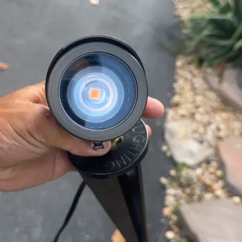 Brass LED landscape light held by hand, showing the bulb's orange and blue glow against a gray surface.