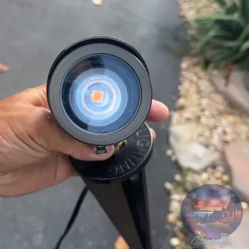 Brass LED landscape light held by hand, showing the bulb's orange and blue glow against a gray surface.