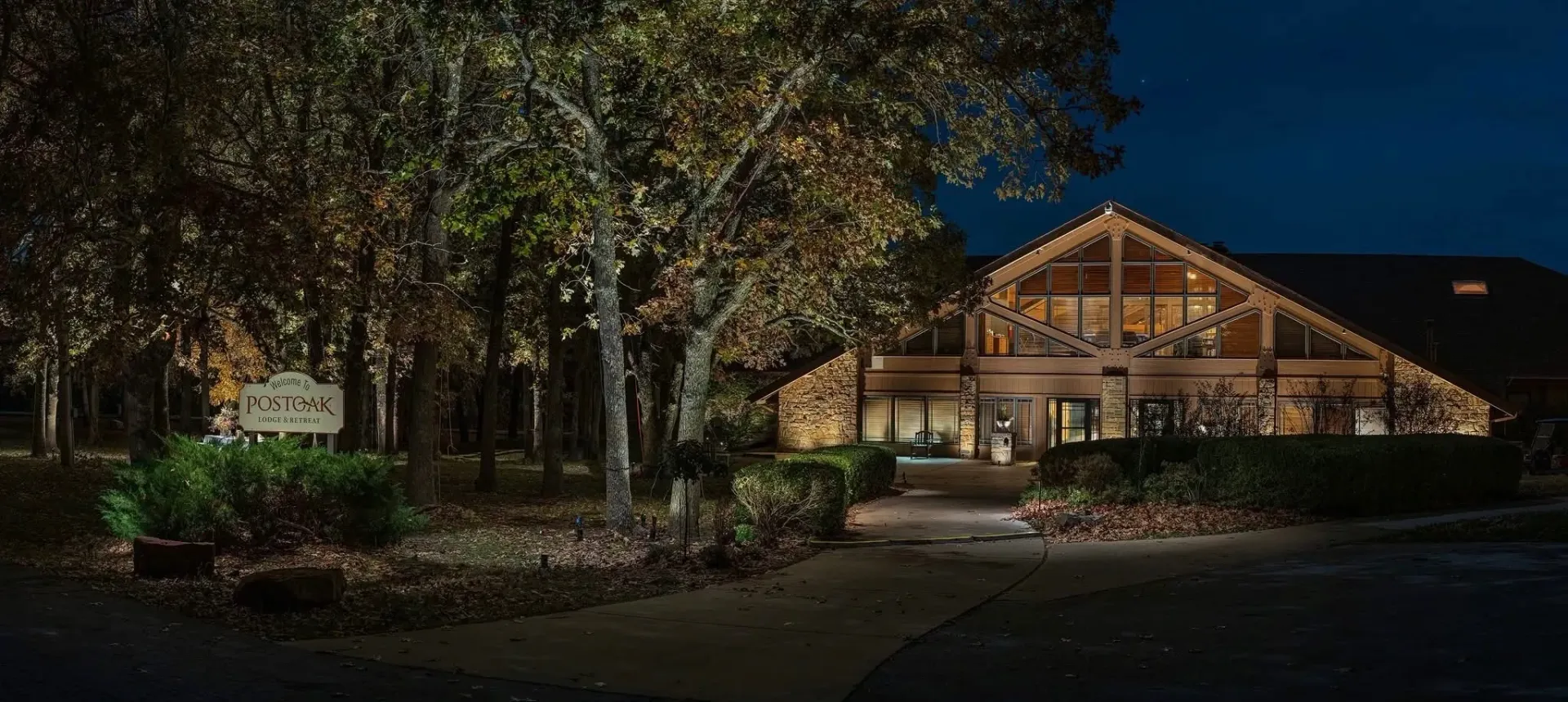 Nighttime view of a wooden lodge with lit windows, surrounded by trees and landscaping.
