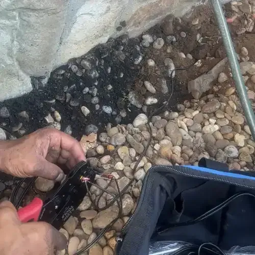 Person using wire strippers on wires near rocks and a black bag.
