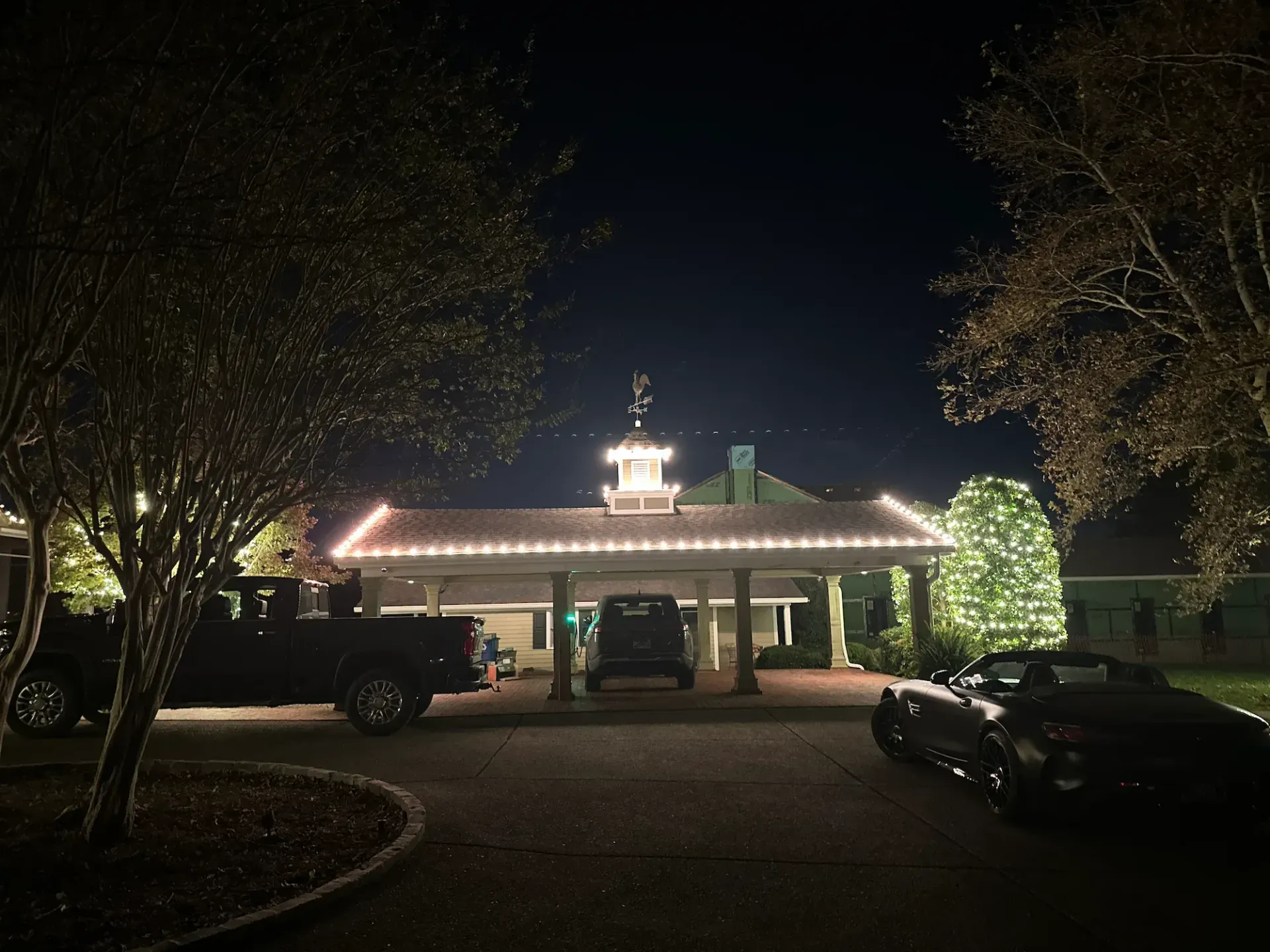 Nighttime view of a building with a decorated entrance lit with Christmas lights; cars parked in the driveway.