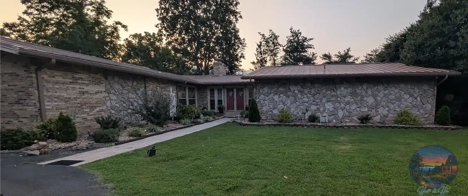 A single-story stone house with a lawn and walkway, trees in the background, and a blue sky.