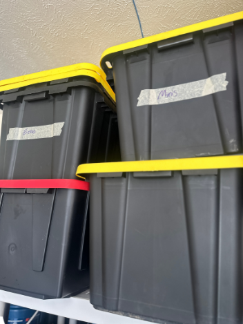 Four black storage bins with colored lids stacked on a shelf. Each bin has handwritten labels.