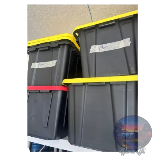 Four black storage bins with colored lids stacked on a shelf. Each bin has handwritten labels.