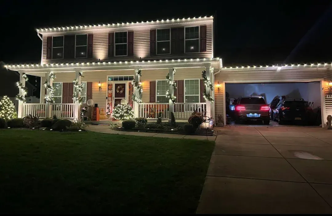 Festive two-story house lit with white Christmas lights; cars in garage; green lawn.