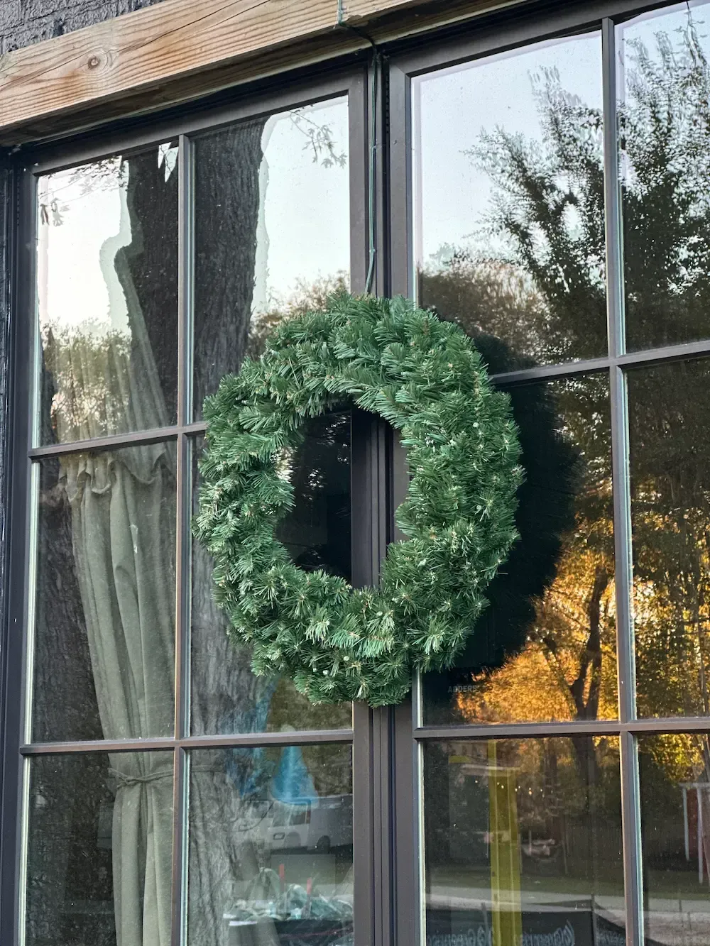 Green leafy wreath on a dark-framed window, reflecting trees and a building.