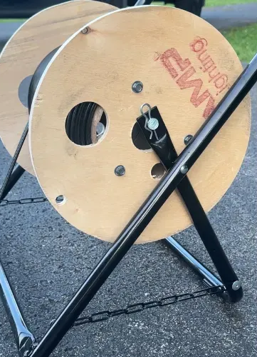 Cable reel with black metal frame, on pavement. Wooden spool has black cable wrapped on it.