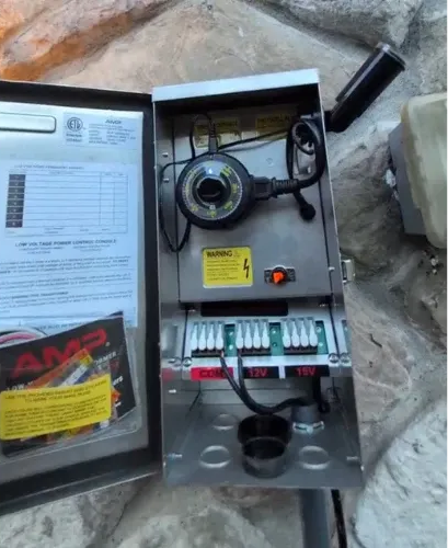 Open electrical box with timer, wiring, and transformer, mounted on a stone wall.