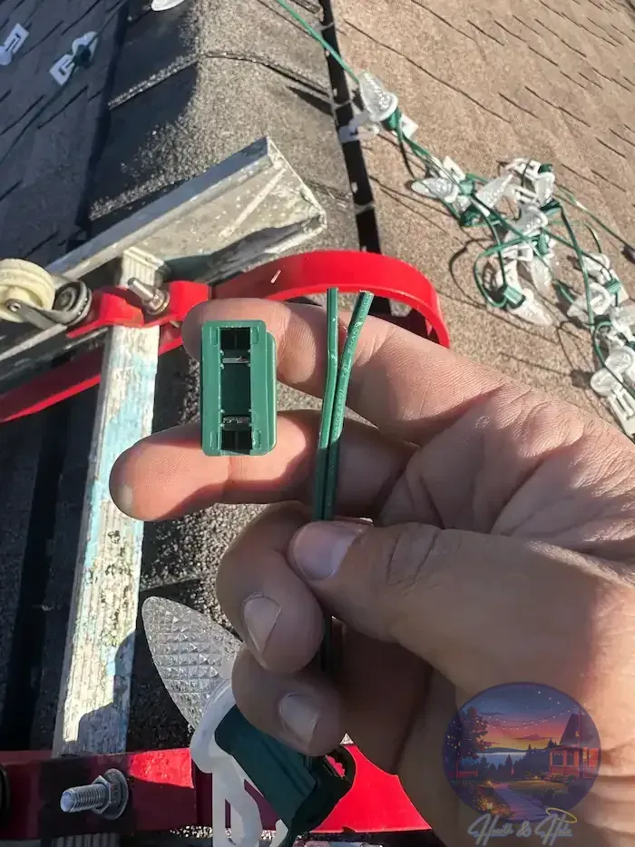 Hand holding a green Christmas light connector with three wires, on a rooftop with lights.