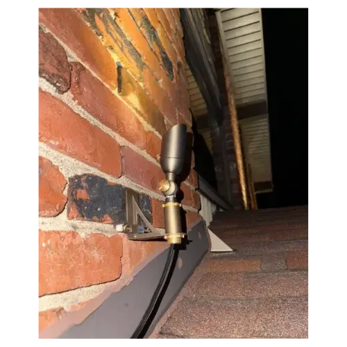A dark bronze spotlight on a brick wall, angled at a roof. The scene is nighttime.