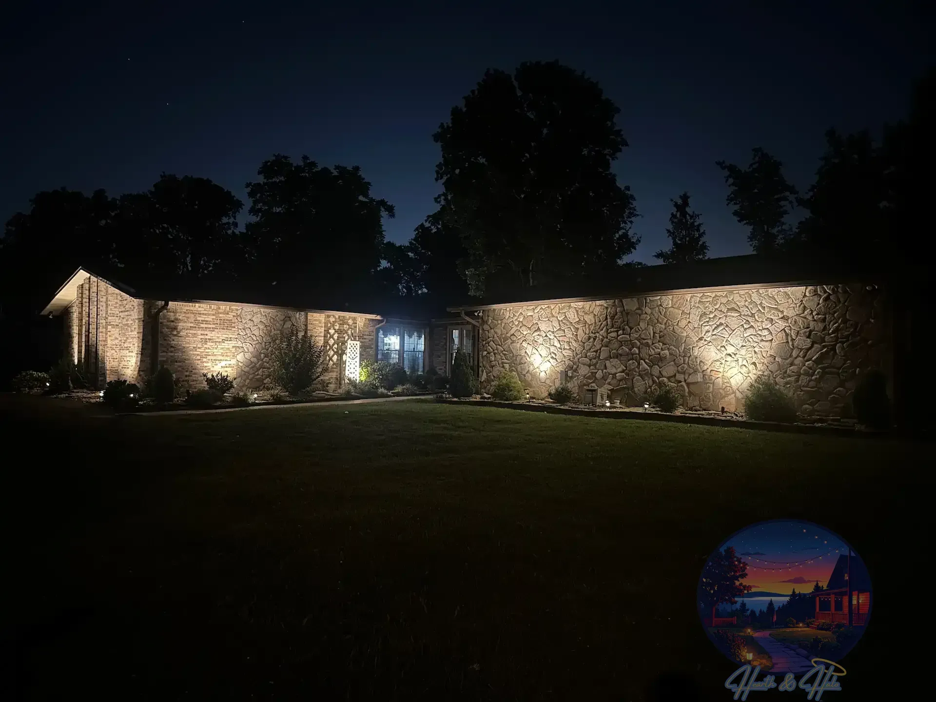 Nighttime view of a stone house with spotlights illuminating the textured walls, set in a grassy yard.