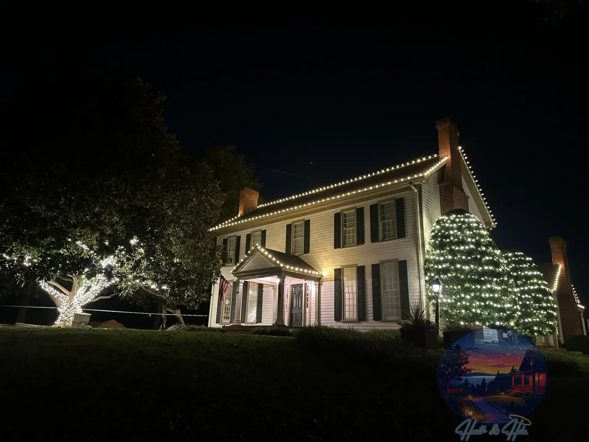 Trees wrapped in string lights illuminate a grassy park at night, with a building in the background.