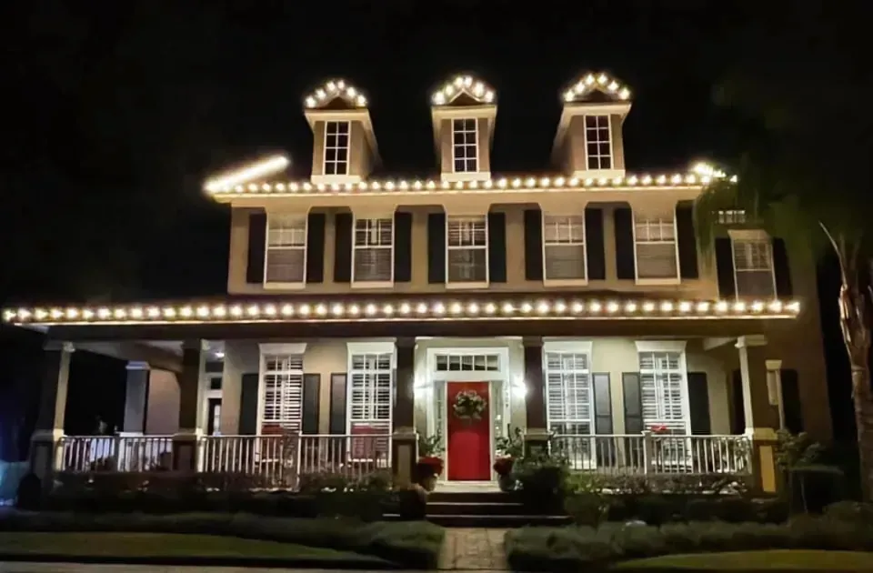 A large two-story house at night, lit with white Christmas lights; red front door, porch with pillars.