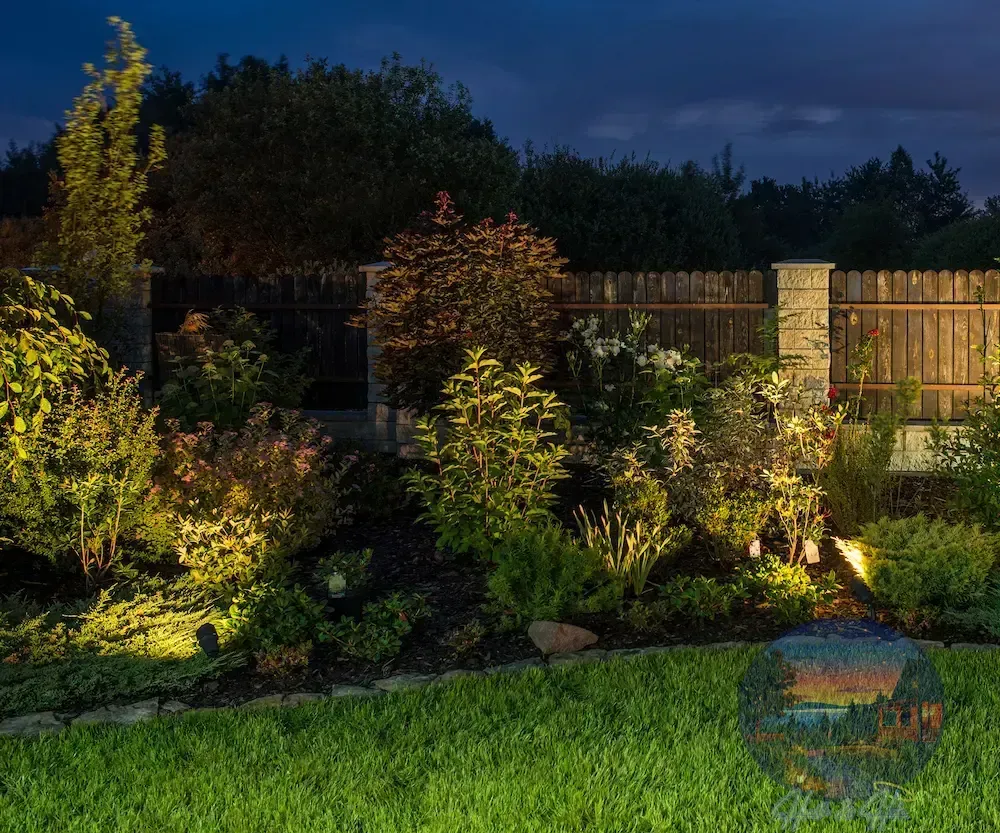 Nighttime garden scene with illuminated plants, wooden fence, and green lawn.