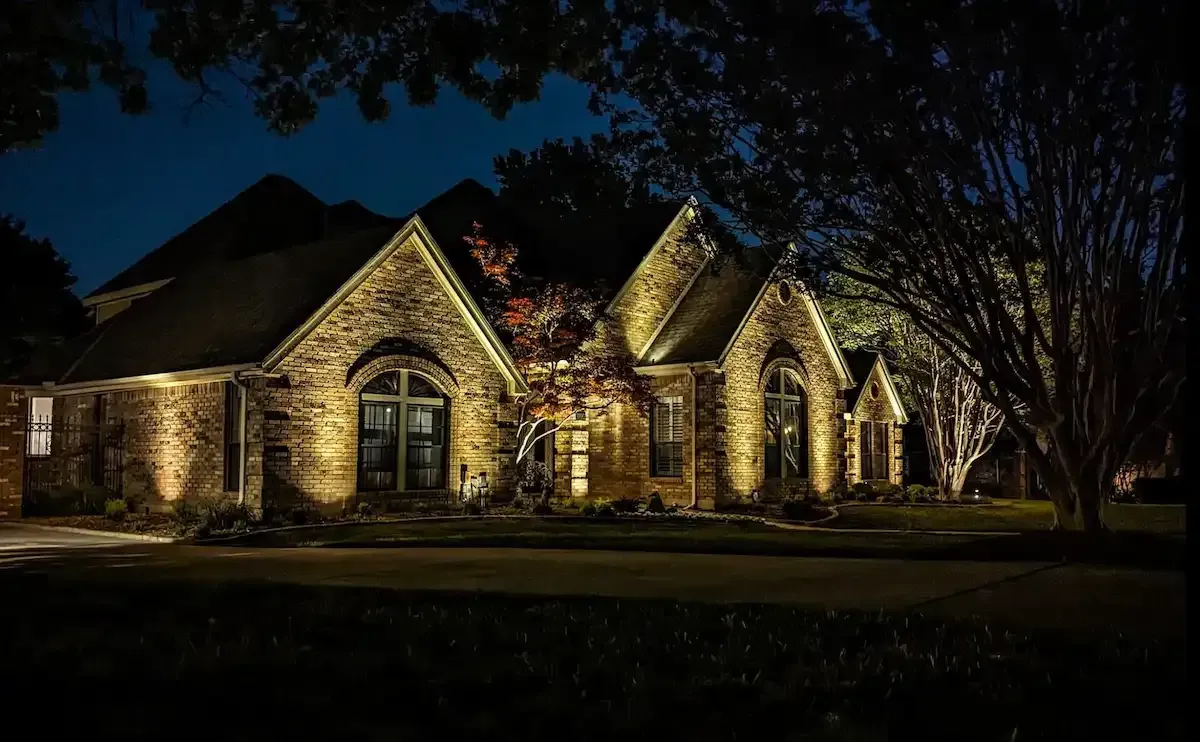 A brick house illuminated at night with warm-toned lights, trees, and a dark sky.