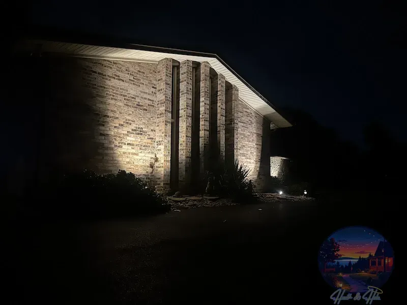 An exterior view of a stone building at night, illuminated by spotlights casting shadows between vertical wall panels.