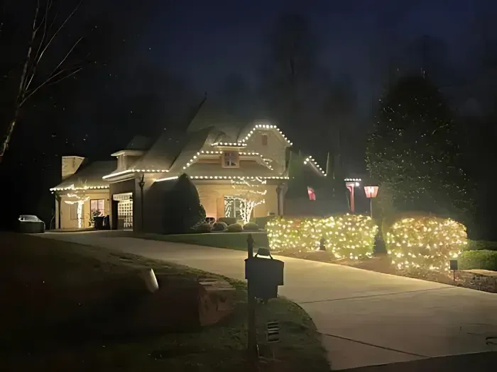 House at night, decorated with white Christmas lights on roof and bushes, driveway.