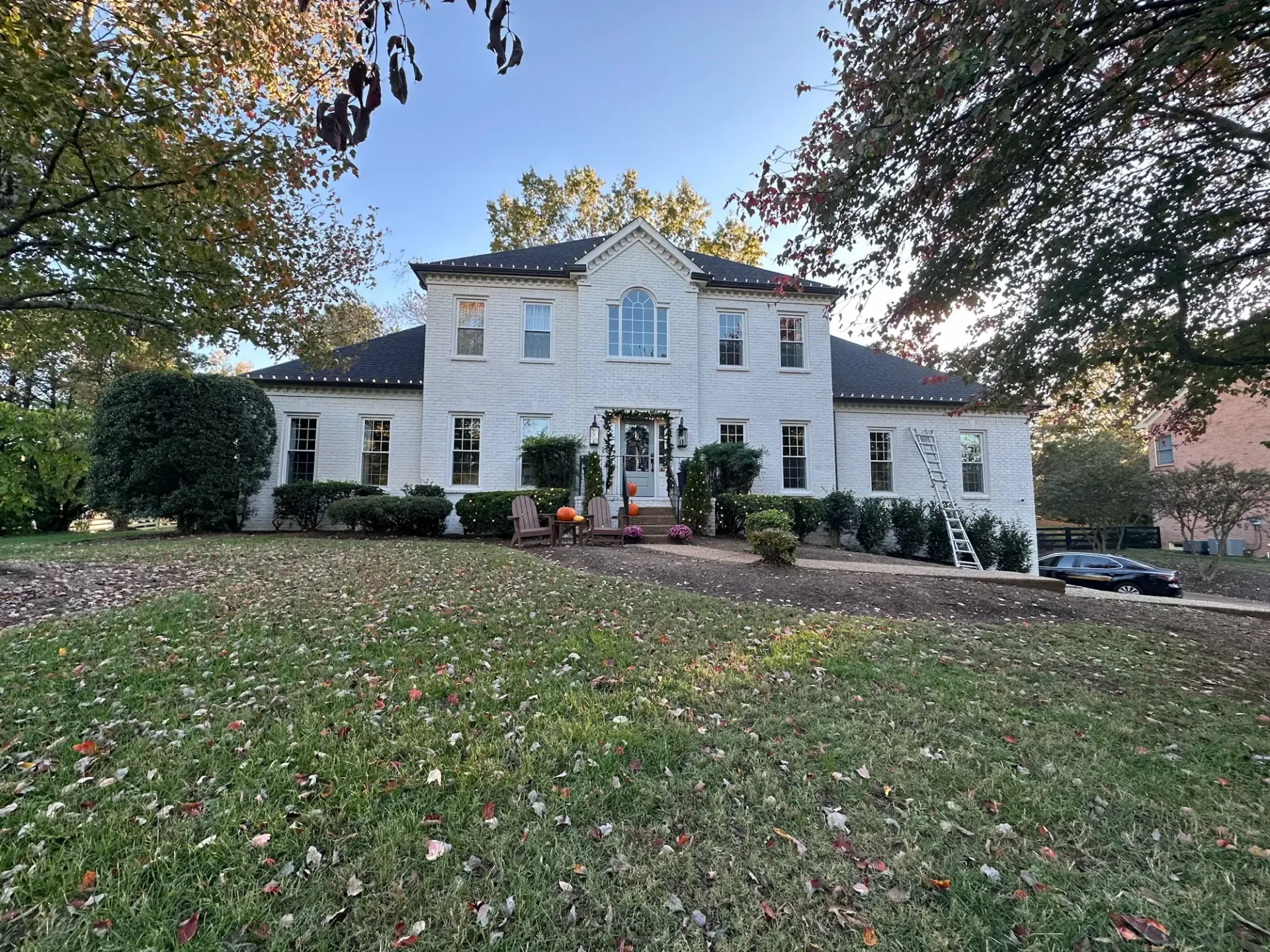 White two-story house with many windows and trees, with a lawn covered in fallen leaves under a blue sky.