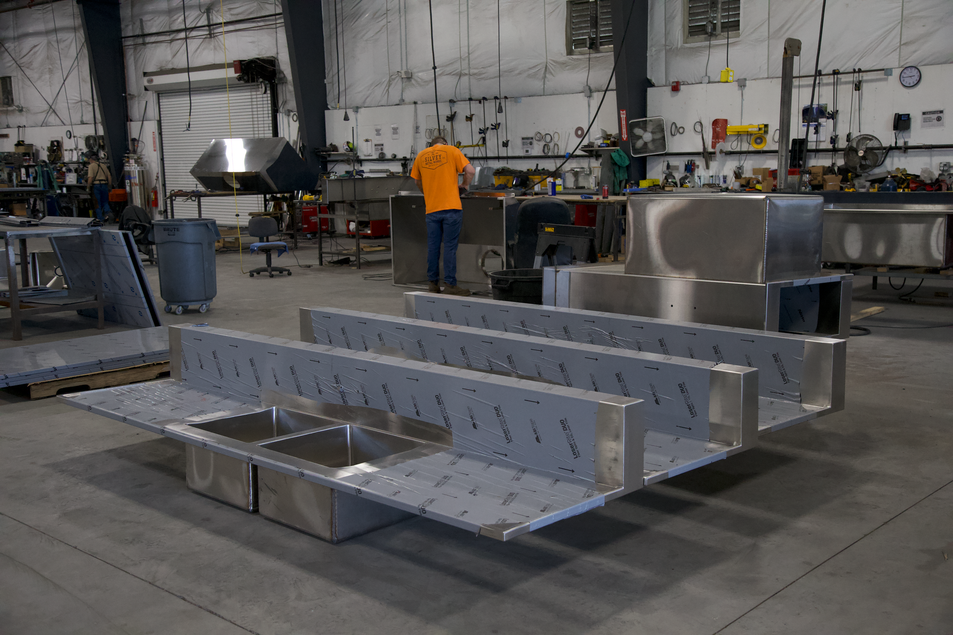A man in an orange shirt is working on a stainless steel sink in a factory.