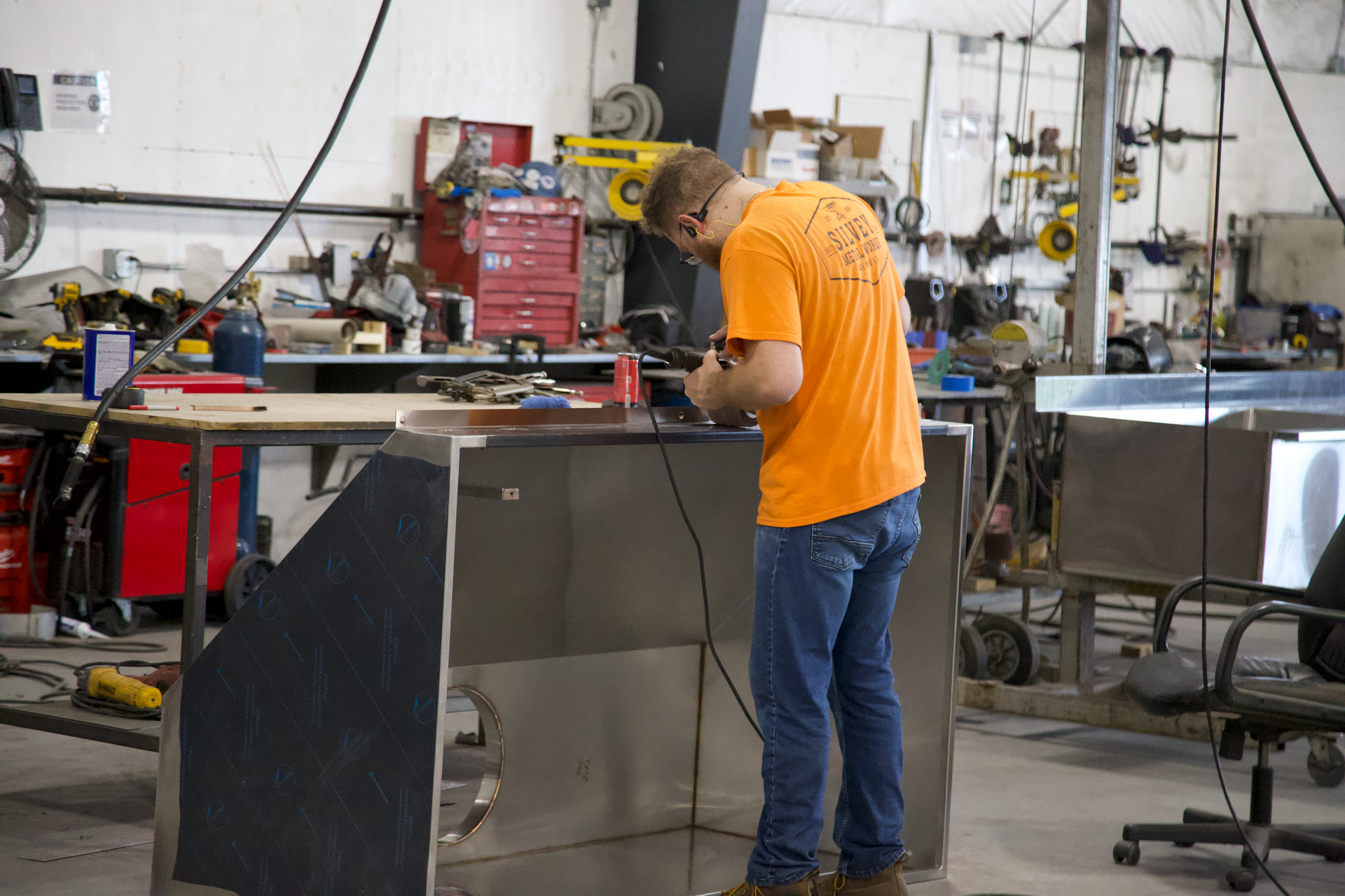 A man in an orange shirt is working on a piece of metal in a workshop.