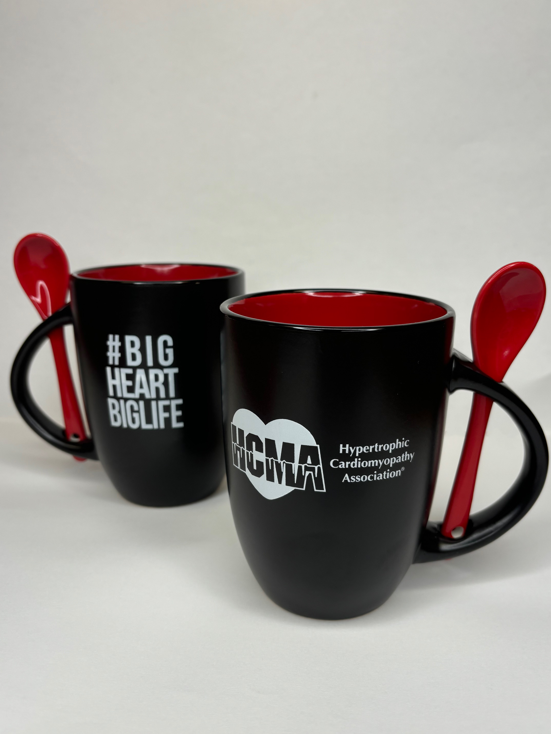 Two black mugs with red interior and a red spoon slotted into the handle of each mug, text #BigHeartBigLife on the mugs