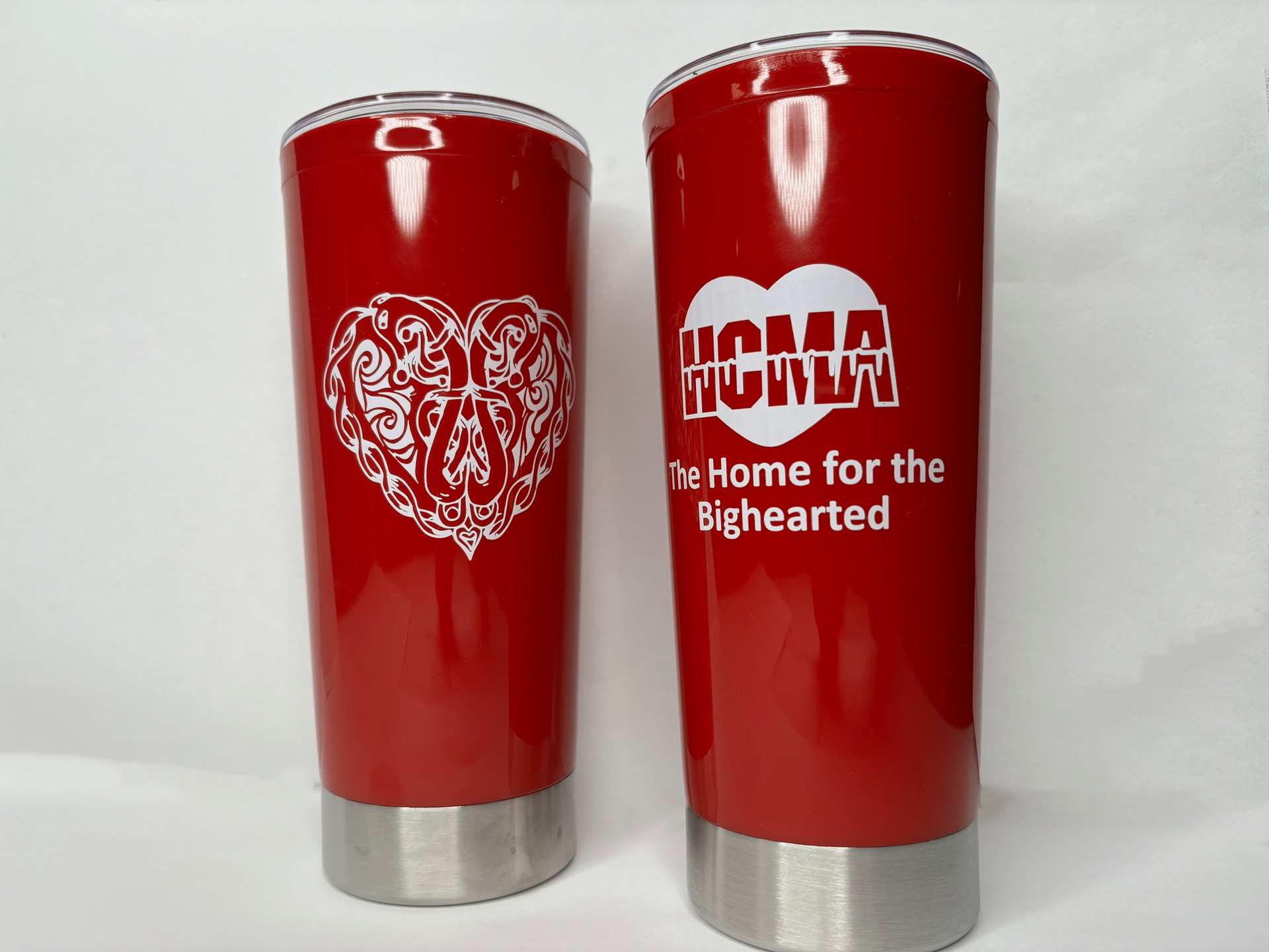 Two red thermal cups with clear plastic tops and silver metal bottoms, showing that one side has a white Celtic heart and the other has the HCMA logo in white with text : The Home for the Bighearted