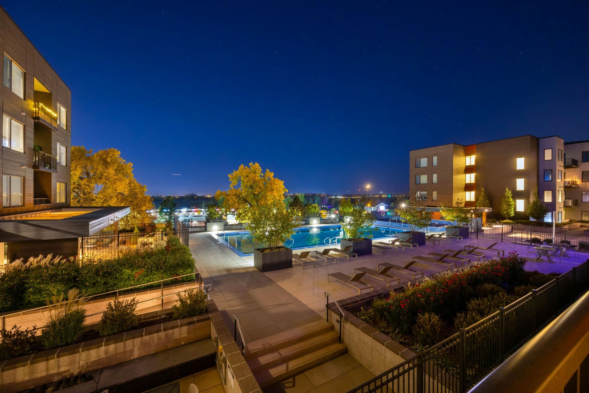 A night view of a brightly lit apartment courtyard featuring a swimming pool surrounded by trees and lounge chairs.