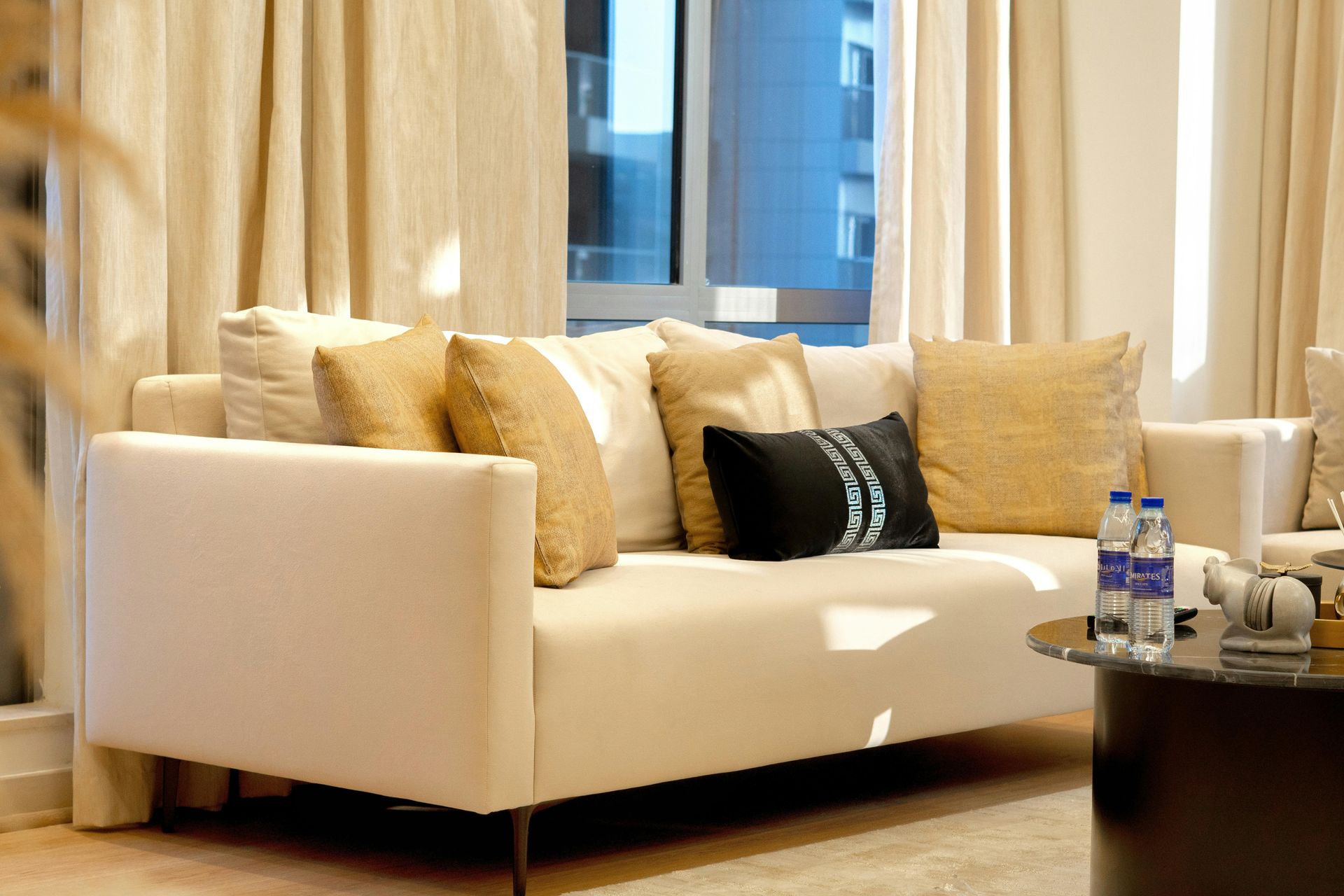 A light-colored sofa with several beige and one black decorative pillow, positioned near a window with floor-length curtains.