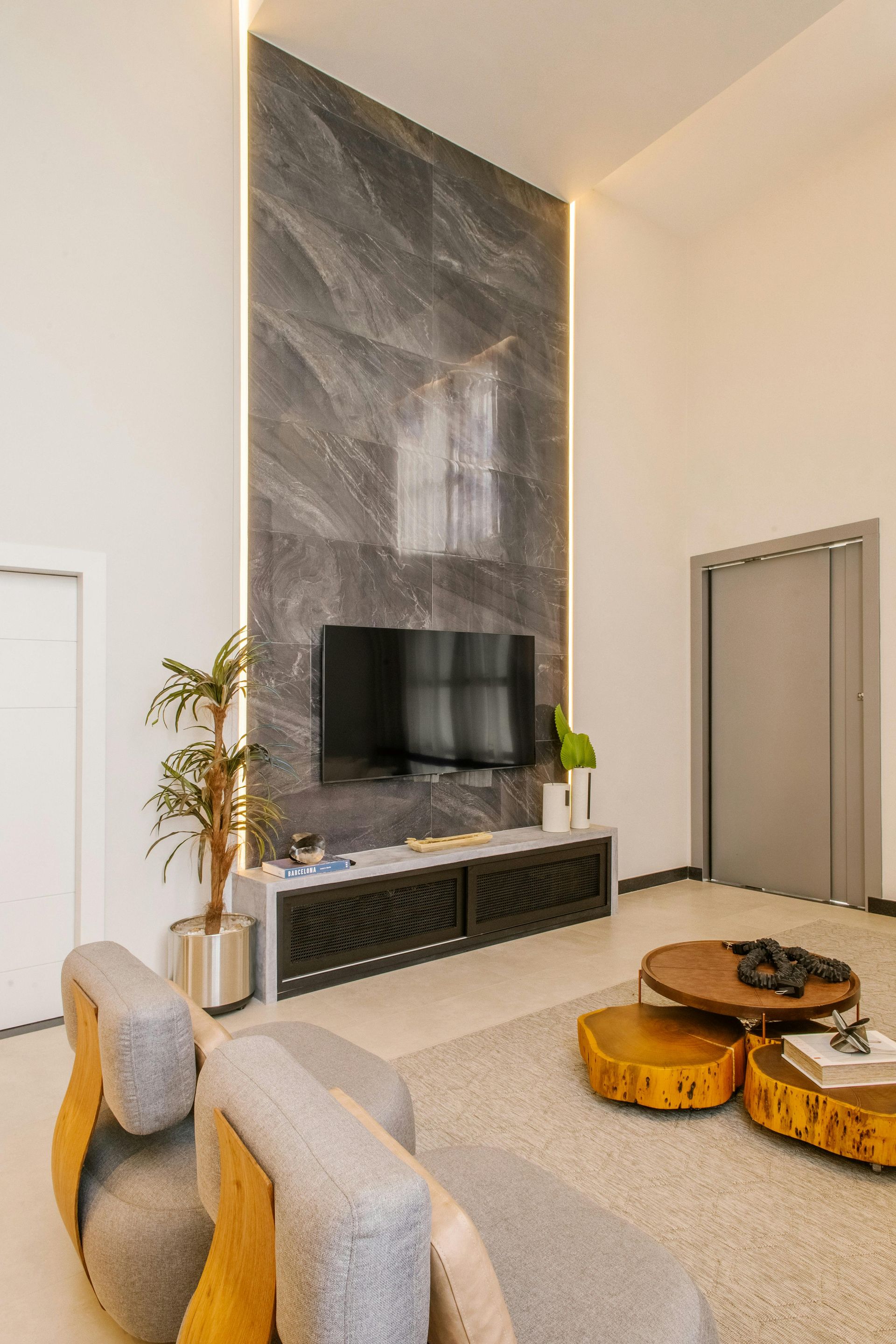 A living room features a dark stone TV accent wall with vertical lighting, a console, a plant, and modern seating.