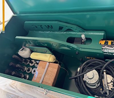 An open green metal storage box containing a hydraulic tool, a coil of cable, metal pins, a cloth, and a wooden block.
