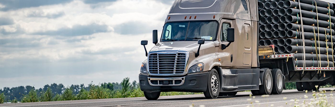 Semi-truck hauling large black pipes on a highway. Cloudy sky, green vegetation on the side.