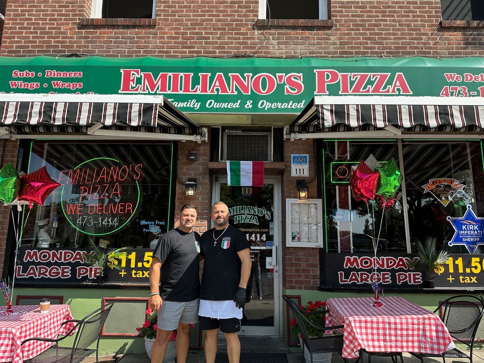 Two men are standing in front of a restaurant called emiliano 's pizza