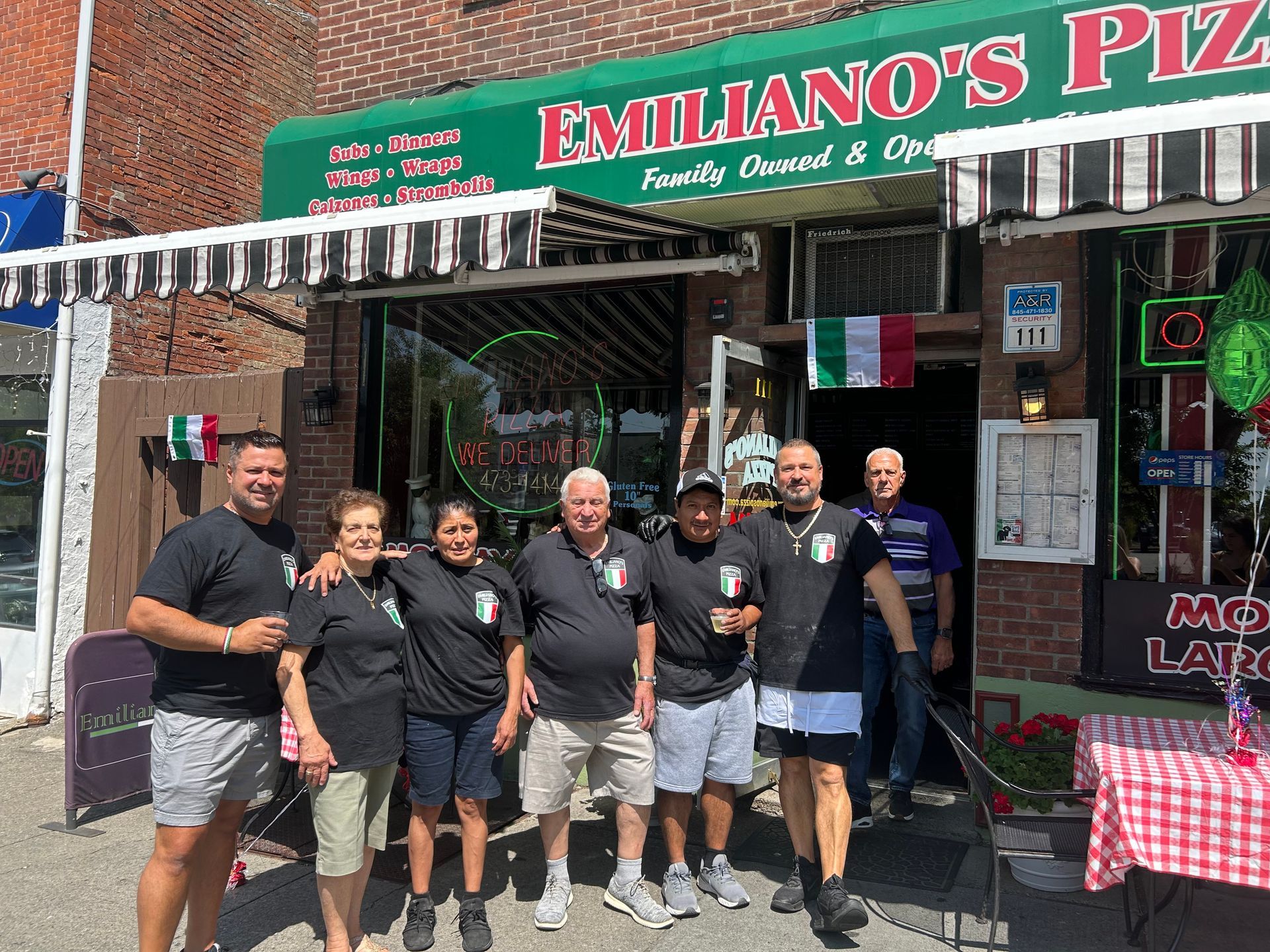 A group of people are posing for a picture in front of a restaurant called emiliano 's pizza.