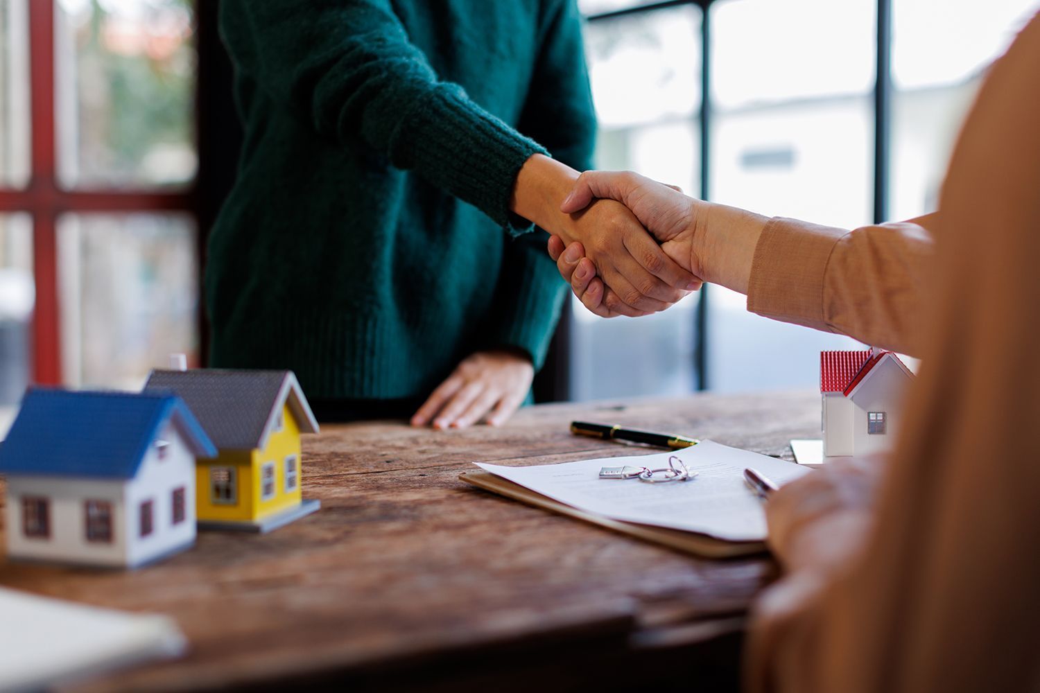 Two people shaking hands over a table with house models and paperwork. Two people shaking hands over a table with house models and paperwork.