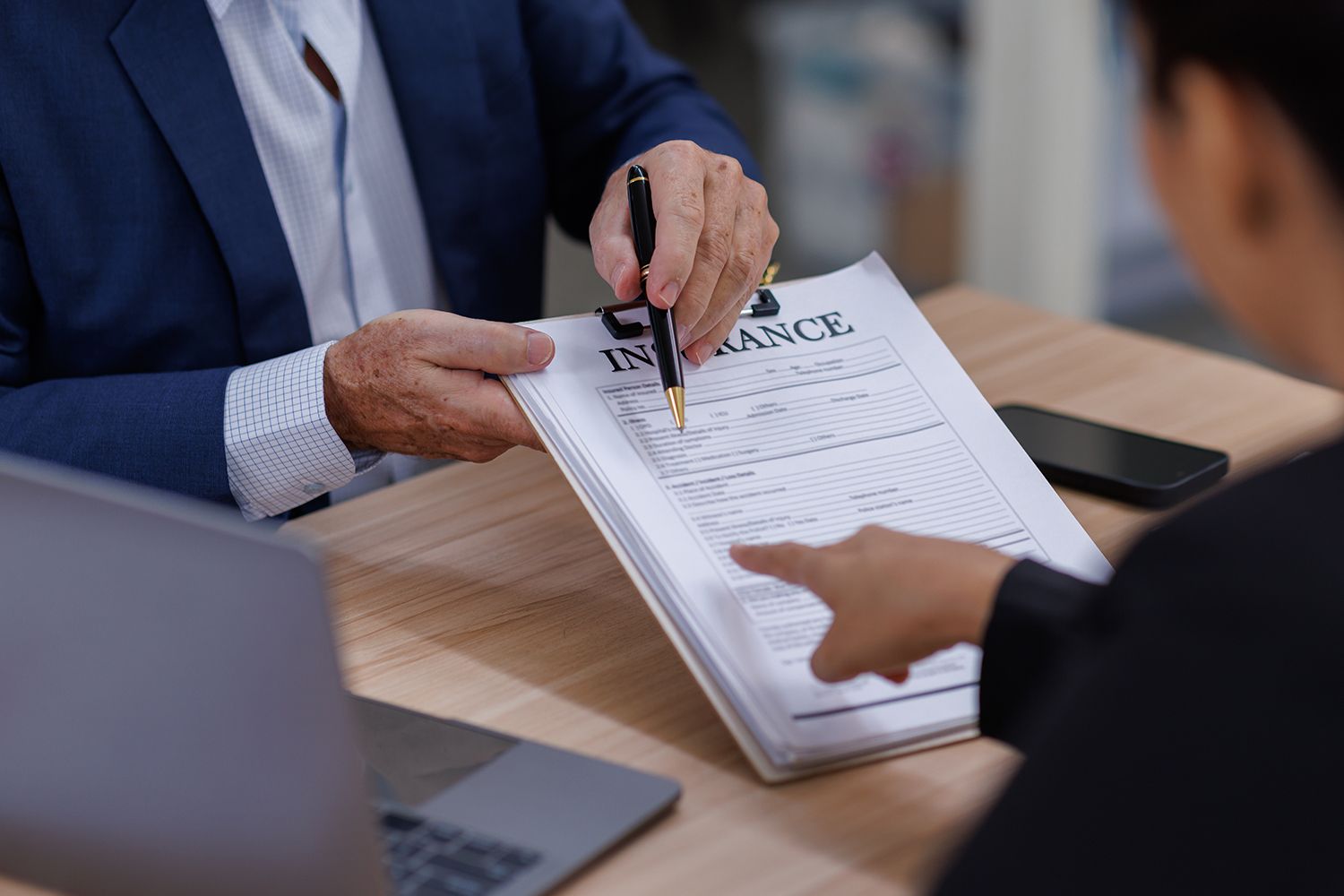 Person points at an insurance document as another person holds a pen, likely signing it. Person points at an insurance document as another person holds a pen, likely signing it.