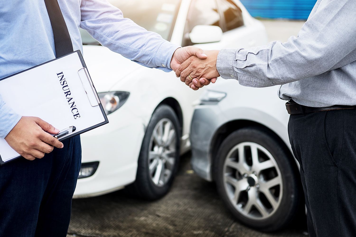 Two men shaking hands beside damaged cars; one holds an insurance form. Two men shaking hands beside damaged cars; one holds an insurance form.