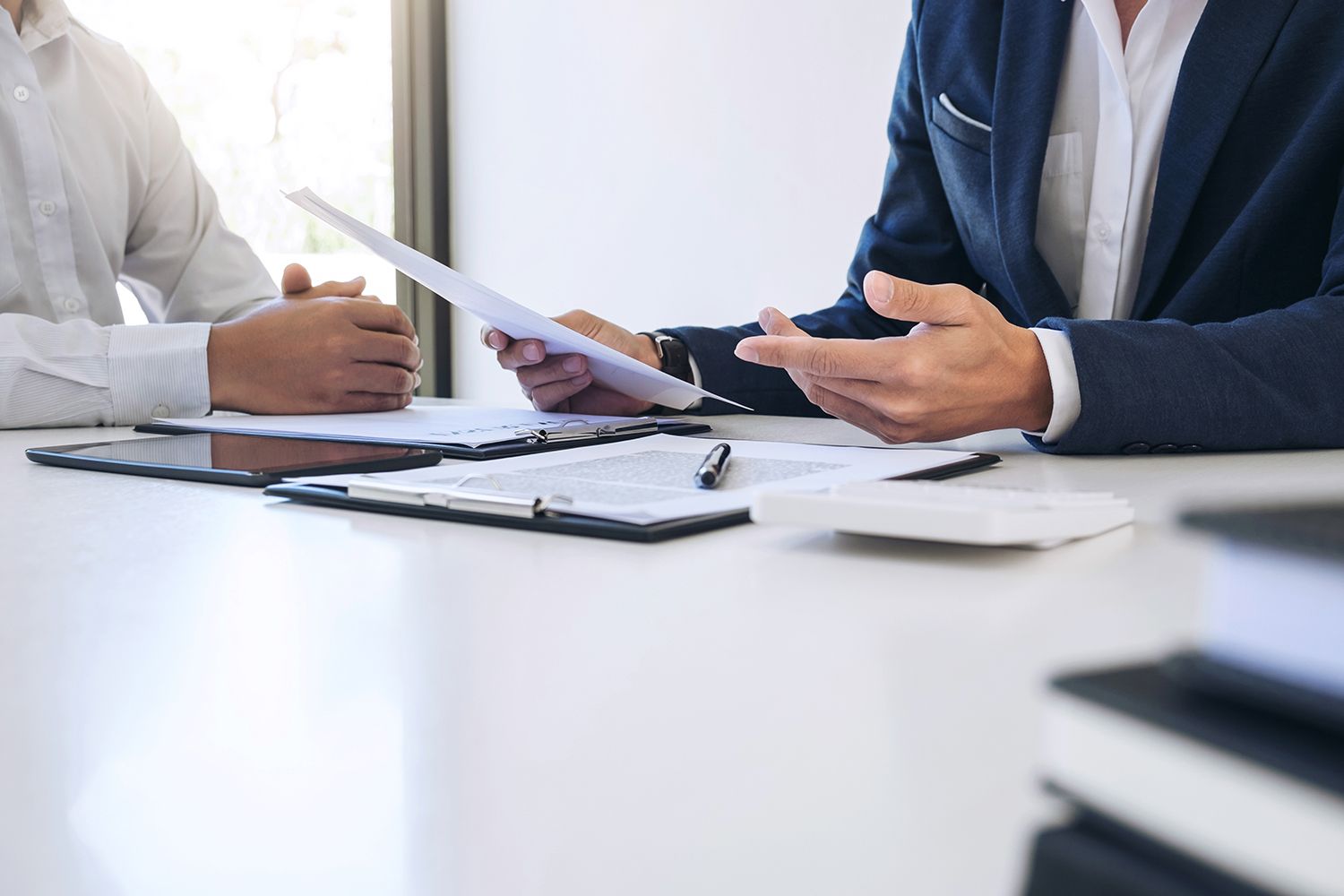 Two men at a desk reviewing documents. One holds a paper, gesturing; the other looks on. Papers and a tablet are on the desk. Two men at a desk reviewing documents. One holds a paper, gesturing; the other looks on. Papers and a tablet are on the desk.