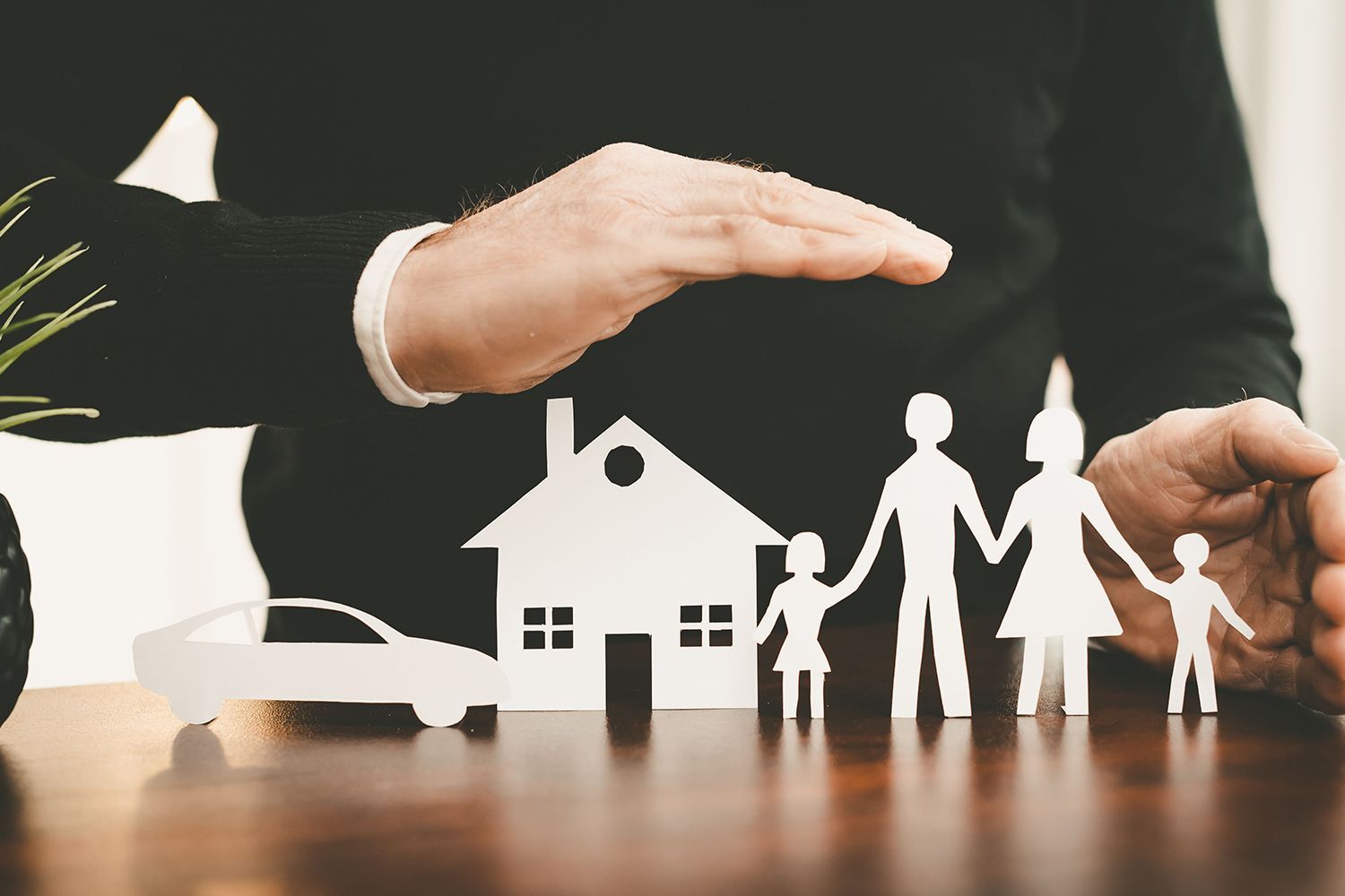 Hand protecting paper cutouts of a family, house, and car on a desk. Hand protecting paper cutouts of a family, house, and car on a desk.