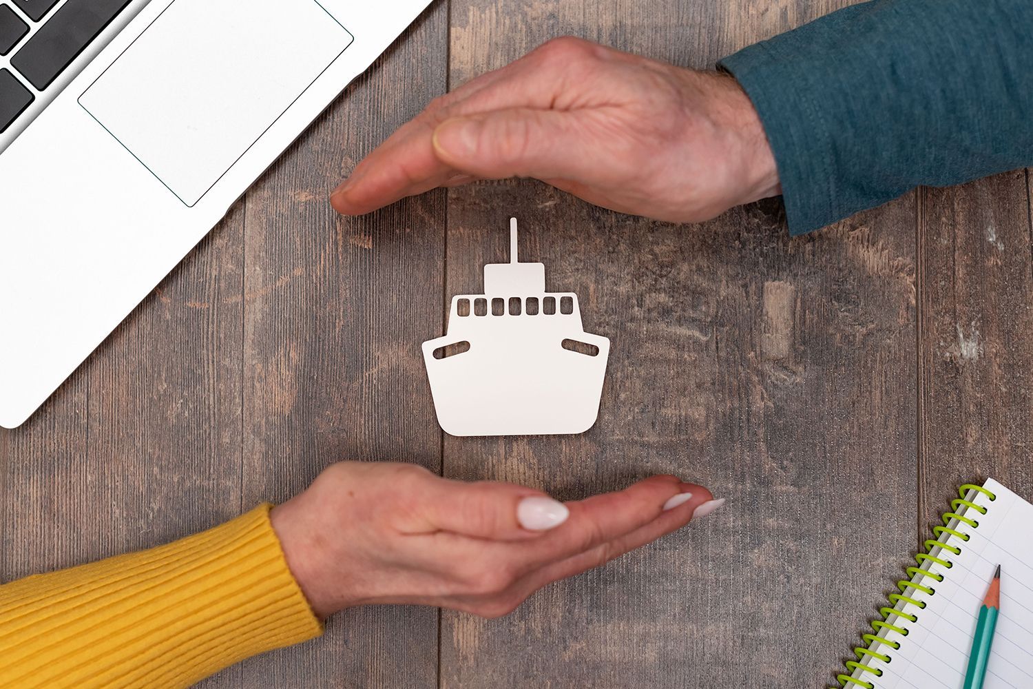 Hands cupping a paper boat over a laptop, notepad, and pen on a wooden surface. Hands cupping a paper boat over a laptop, notepad, and pen on a wooden surface.