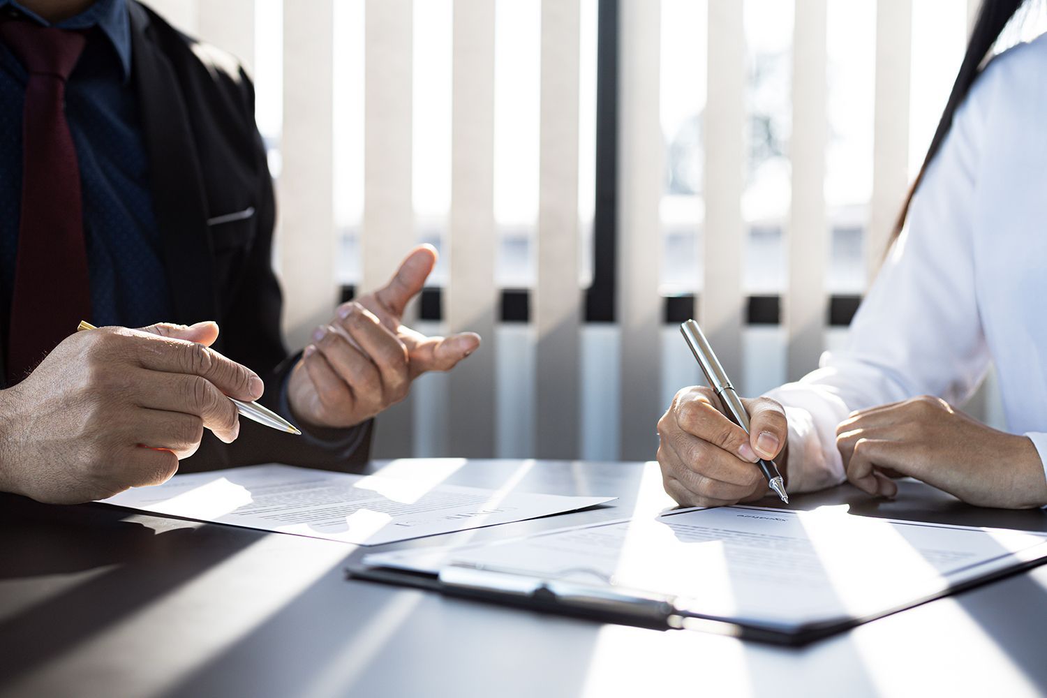 Person in suit gestures, while another signs document at table. Sunlight streams through blinds. Person in suit gestures, while another signs document at table. Sunlight streams through blinds.