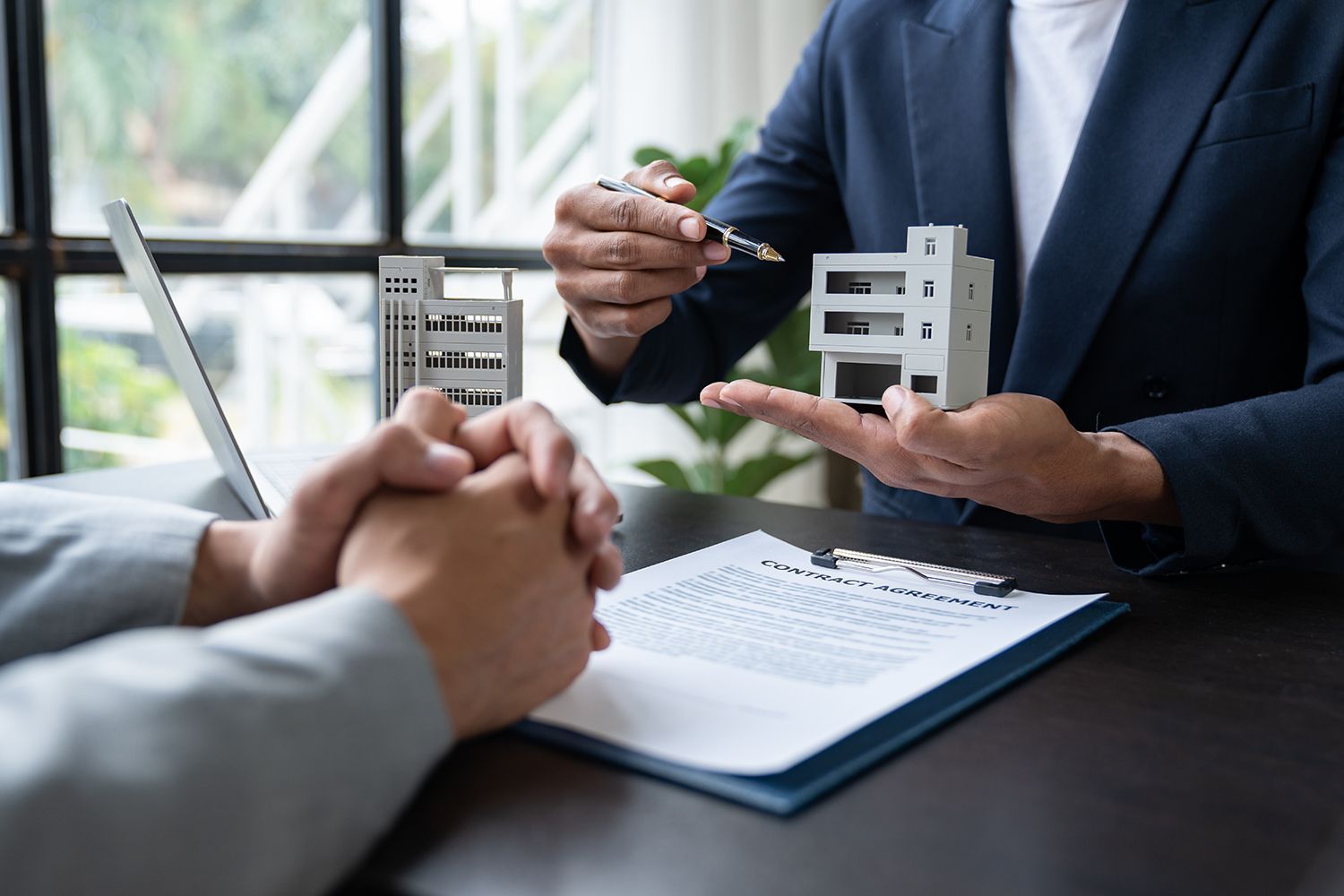 Real estate agent pointing at a model building, presenting a contract. Real estate agent pointing at a model building, presenting a contract.