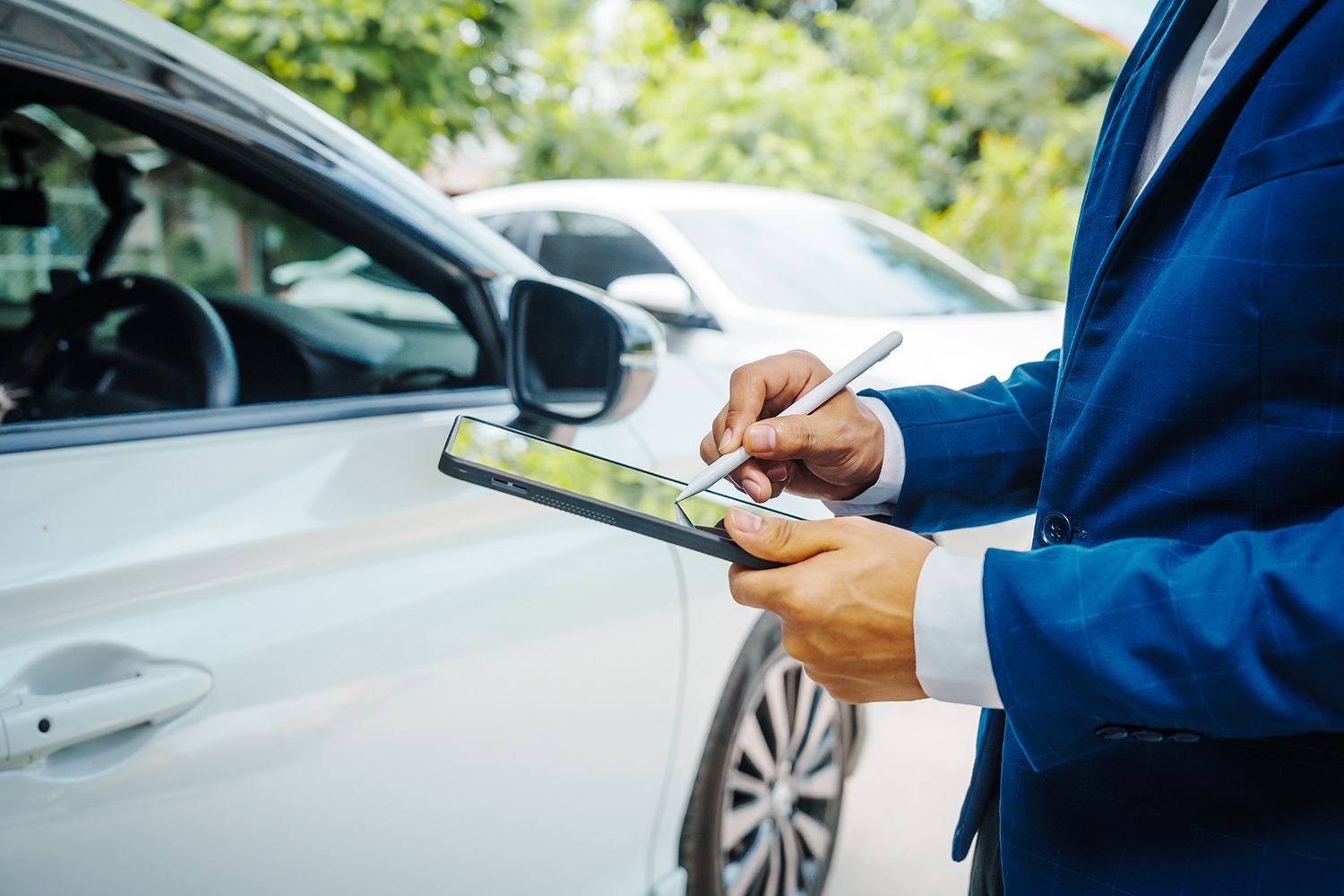 Person in a blue blazer using a tablet near a white car, possibly assessing damage or documenting information. Person in a blue blazer using a tablet near a white car, possibly assessing damage or documenting information.