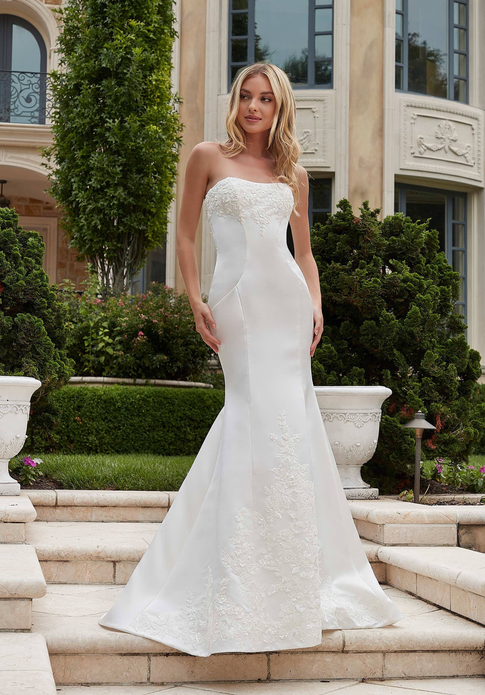 Our Phylicia designer wedding dress is a modern fit and flare with romantic touches. The chic gown glows in Duchess satin with contoured seaming to enhance your curves. The straight strapless neckline is accented by delicately beaded floral embroidery as well as the hemline to create a beautiful border on the chapel length train. Pair the dress with the matching Duchess Satin Overskirt with Floral Beaded Embroidery, sold separately as Style 11524, for a fuller silhouette. Shown in Ivory. Wedding dress by Morilee.